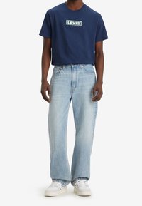 Granatowy bawełniany T-shirt z logo "LEVI'S", zestawiony z jasnoniebieskimi jeansami. Model nosi białe sneakersy. Luźny krój i swobodna sylwetka.