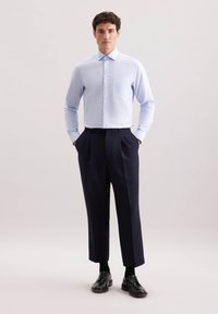 Camicia azzurro chiaro a maniche lunghe con taschino frontale, abbinata a pantaloni blu navy con pieghe e scarpe eleganti nere, presentata su uno sfondo neutro.
