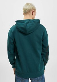 Teal hettegenser med snøring i hetta, utstyrt med kenguru-lomme og et stort trykt grafisk motiv på ermet. Mykt stoff med ribbestrikkede mansjetter.