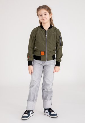 Olivgrüne Bomberjacke mit Reißverschluss, Fronttaschen und orangefarbenem Etikett. Kombiniert mit hellgrauen Jeans und schwarzen Turnschuhen.
