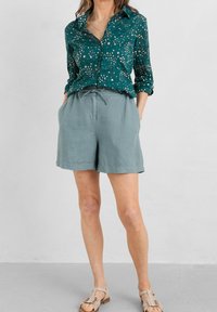 Blouse à motifs bleu sarcelle à manches longues, associée à un short en lin gris clair. Sandales plates complètent la tenue, avec une coupe décontractée et une taille à cordon.