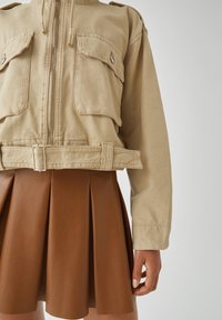Jasno brązowa, krótka kurtka z zamkiem błyskawicznym, dwiema kieszeniami na piersi oraz paskiem w talii, noszona na brązowej plisowanej spódnicy z ekologicznej skóry.