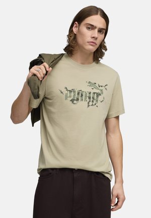 Camiseta verde oliva claro de algodón con un logo gráfico estilo camuflaje. El modelo sostiene una chaqueta verde oscuro sobre su hombro.