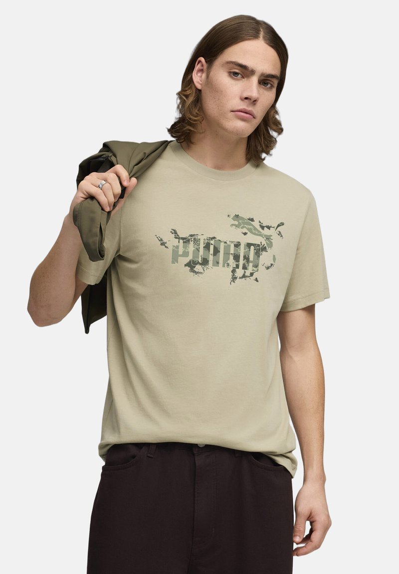 T-shirt vert olive clair en coton avec un logo graphique style camouflage. Le modèle porte une veste vert foncé sur l'épaule.
