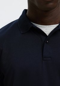 Uomo che indossa una polo blu scuro su una t-shirt grigio chiaro con scollo tondo, mostrando la linea del collo e la parte superiore del petto.