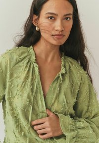 Zielona, teksturowana bluzka o luźnym kroju, z postrzępionymi detalami, dekoltem w kształcie litery V i długimi rękawami, z wiązaniem przy dekolcie.