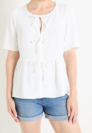 Vrouw draagt een witte blouse met korte mouwen met drie gestrikte strikken aan de voorkant en blauwe opgerolde spijkershorts.