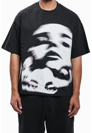 Homme portant un t-shirt noir oversize avec une grande impression abstraite d'un visage flou en noir et blanc sur le devant.
