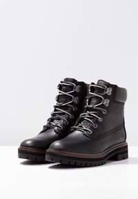 Bottes noires en cuir à lacets avec ferrures argentées, côtés texturés et semelles robustes, avec des lacets gris et des semelles intermédiaires marron contrastantes.