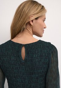 Tumma vihreä mekko, jossa on smokkitekstuuri, pitkät läpinäkyvät hihat ja avausyksityiskohta takana kaula-aukossa. Sileä kangaspinta.