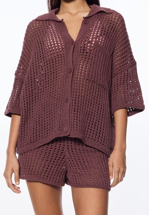 Femme portant un ensemble violet assorti composé d'un haut à boutons ample en maille ajourée avec poche et d'un short, mettant en valeur le motif texturé et la coupe décontractée.