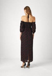 Frau steht seitlich mit freiem Rücken, trägt ein schwarzes, schulterfreies Maxikleid mit rotem Blumenmuster und hohe Sandalen.