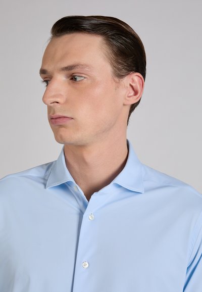 Camisa azul claro con botones, cuello clásico, textura suave y brillo sutil. Presenta botones blancos y detalles de costura impecables.