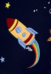 Razzo ricamato giallo e rosso con finestre blu e scia arcobaleno su tessuto blu scuro con stelle ricamate.