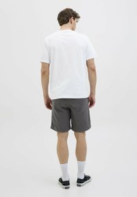 Camiseta de algodón blanca, con cuello redondo, combinada con pantalones cortos grises. El modelo lleva calcetines blancos de caña corta y zapatillas negras, visto desde la parte de atrás.