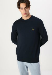 Maglione lavorato a maglia blu navy con scollo rotondo, maniche lunghe, orlo aderente e un piccolo logo di un uccellino giallo sul petto. Tessuto liscio.