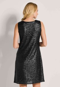 Frau mit blonden Haaren, die ein ärmelloses schwarzes Paillettenkleid trägt, von hinten vor einem schlichten hellen Hintergrund gezeigt.