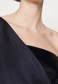 Černé saténové šaty s nařaseným, asymetrickým výstřihem a hladkou texturou. Oděv má hluboký tvar do V na rameni.