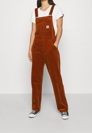Manchesteroveralls i brændt orange med justerbare stropper, brystlomme og sidelommer, stylet med en hvid T-shirt og sorte sneakers.