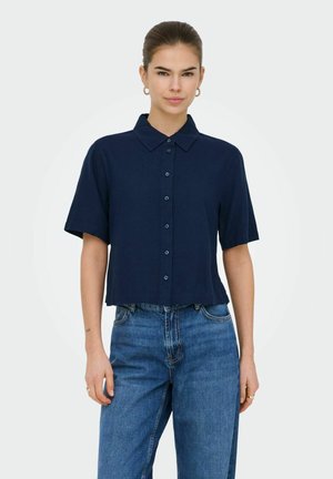 Donna con capelli scuri raccolti in uno chignon, orecchini a cerchio dorati, camicia blu navy a maniche corte con bottoni e jeans blu a vita alta su sfondo bianco.