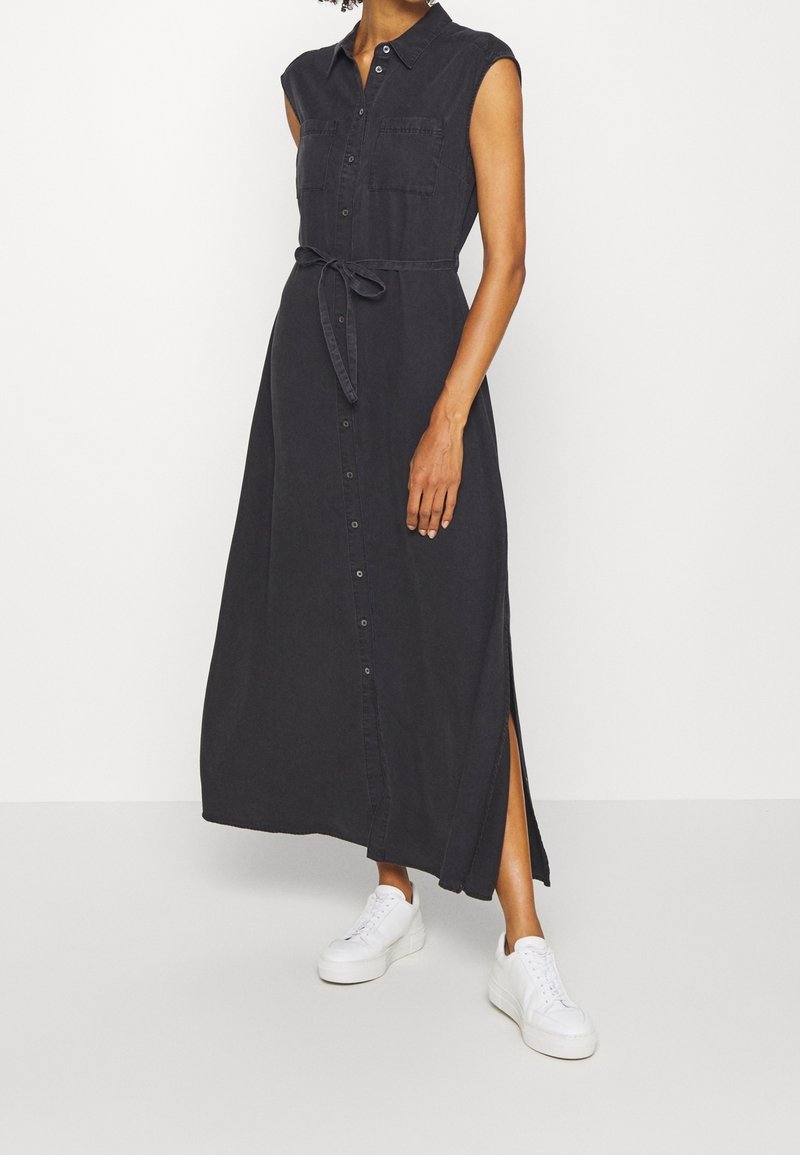Robe maxi sans manches noire avec un devant à boutons, deux poches poitrine, taille ceinturée et fentes sur les côtés, associée à des baskets blanches.