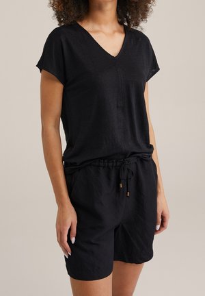 Kobieta ubrana w czarną bluzkę z krótkim rękawem i dekoltem w serek oraz czarne szorty ze sznurkiem, na neutralnym tle.