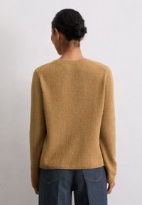 Mustard yellow textured sweater with long sleeves and a round neckline, paired with dark denim pants. Back view emphasizes the soft fabric.