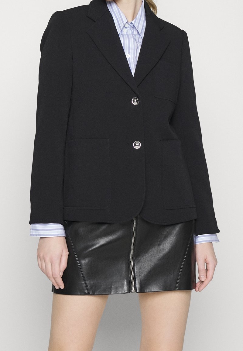 Femme portant un blazer noir sur une chemise rayée bleue, associée à une mini-jupe en cuir noire avec une fermeture éclair à l'avant.