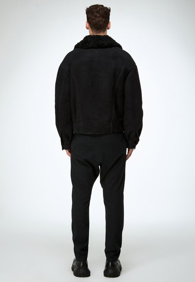 Veste en daim noire avec un col en shearling noir texturé, associée à un pantalon noir et des chaussures noires épaisses, vue de dos.