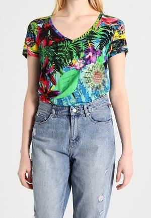 Vrouw draagt een kleurrijk, bloemig, kort mouw V-hals top, ingestopt in een lichtblauwe, hoog getailleerde, versleten spijkerbroek.