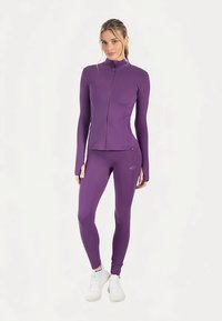 Veste de sport violette avec col haut et coupe ajustée, associée à un legging assorti. Les deux sont fabriqués en tissu extensible. Baskets blanches.