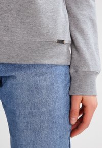 Sudadera gris con puños y dobladillo acanalados, con una pequeña etiqueta negra con detalles en naranja. Combinada con jeans de mezclilla azul.