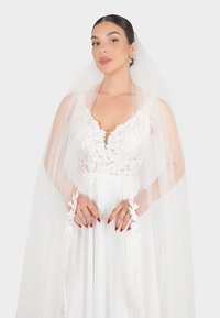 Abito da sposa bianco con dettagli in pizzo floreale e scollatura a V profonda, abbinato a un lungo velo trasparente, indossato su una superficie di tessuto liscio.