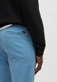 Pantalons bleu clair en tissu lisse, dotés d'une poche arrière avec une étiquette "BOSS" noire et rouge, portés avec un sweat-shirt noir.