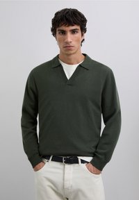 Suéter tejido verde con escote en V y cuello vuelto, de mangas largas, combinado con una camisa blanca interior y pantalones beige, que cuentan con un cinturón negro.