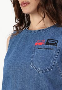 Top de denim sem mangas com gola redonda, um bolso no peito com o texto "KARL LAGERFELD" e detalhes gráficos em bordado nas cores vermelho e preto.