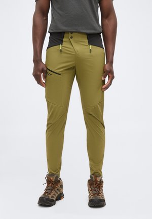 Mand iført olivengrønne vandrebukser med sorte taljepaneler og brune vandrestøvler, stående mod en lys baggrund.