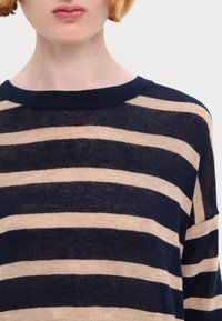 Person in einem navy- und beige gestreiften Strickpullover, von Hals bis zur oberen Brust gezeigt, mit heller Haut und kurzen hellen Haaren.