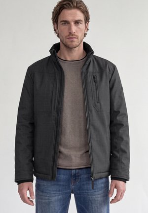 Mand med lyst brunt hår iført en sort lynlåsjakke over en brun skjorte og blå jeans, stående mod en ensfarvet lys baggrund.