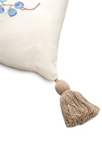 Αφράτος μαξιλάρι σε κρεμ απόχρωση με υφή, μπλε κεντητές μούρες και μια μεγάλη, μπεζ φούντα ως αξεσουάρ σε μια γωνία.