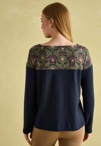 Marineblaue Langarmbluse mit einem floral gemusterten Einsatz in den Farben Rot, Grün und Creme; glatte Textur; lockere Passform, weiter Halsöffnung.