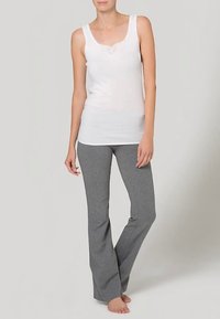 Sieviete stāv basām kājām, ģērbusies baltā bez piedurknēm tops un pelēkās pieguļošās jogas biksēs uz vienkārša fona.