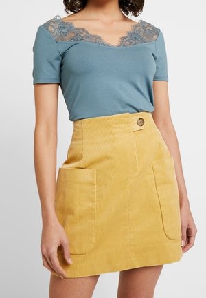 Femme portant un haut bleu à manches courtes avec un col en dentelle et une jupe en velours côtelé jaune avec de grandes poches avant et un détail de boutons.