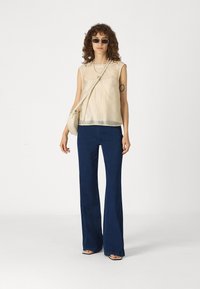 Beige sleeveless top with sheer hem, paired with dark blue flared jeans. Model accessorized with sunglasses and a small crossbody bag.