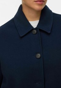 Približen posnetek osebe, ki nosi mornarsko moder volnen plašč z velikim ovratnikom in črnimi gumbi preko bele srajce.