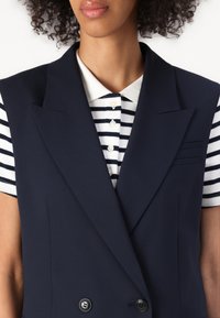 Marineblå ermeløs blazer med dobbeltradet design, som har slag og to frontlommer over en rutete polo-skjorte.