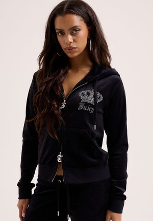 Frau trägt schwarzen Velours-Kapuzenpullover mit Reißverschluss, verziert mit einer Strass-Krone und dem "Juicy"-Logo, kombiniert mit passenden schwarzen Hosen, vor einfarbigem Hintergrund.