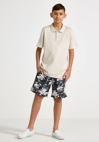 Beige kortärmad pikétröja med tre knappar, tillsammans med marinblå badshorts med vit palmträdstryck, och vita slip-on skor.