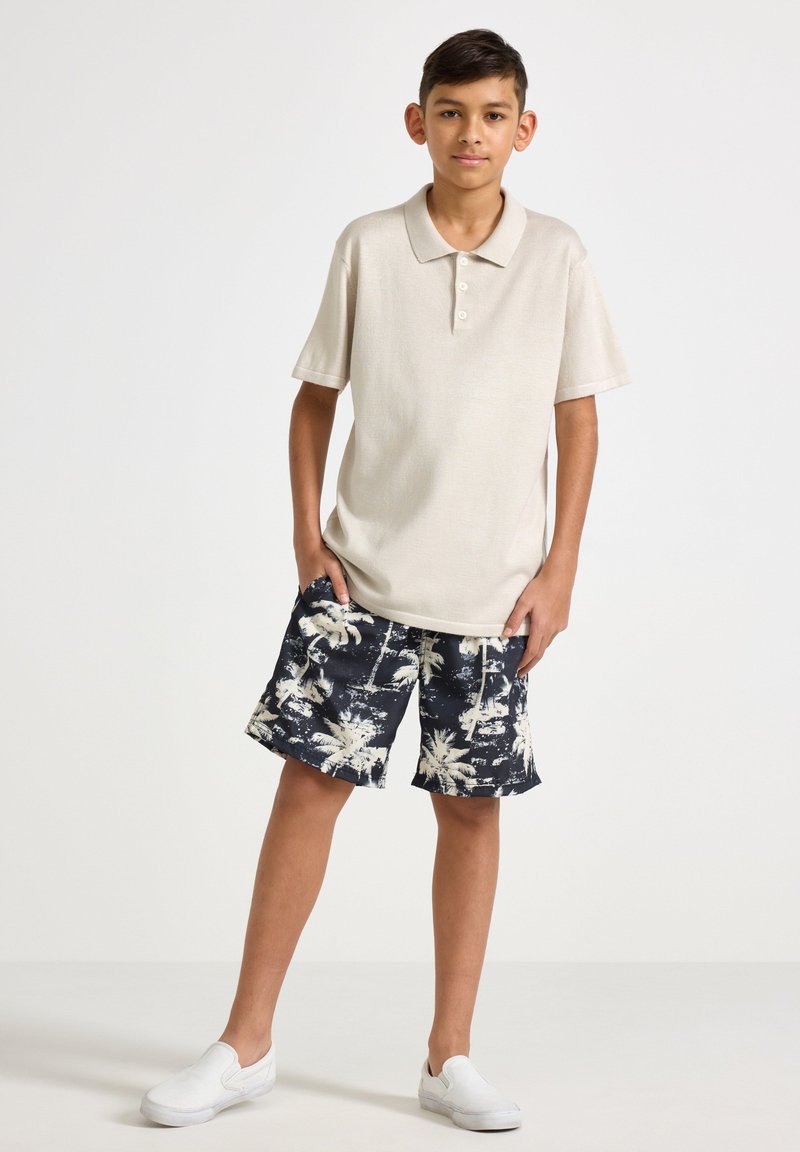 Beige kortärmad pikétröja med tre knappar, tillsammans med marinblå badshorts med vit palmträdstryck, och vita slip-on skor.