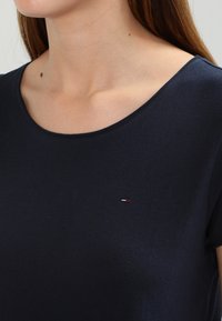 Žena v námořnickém modrém tričku s kulatým výstřihem a krátkým rukávem, na kterém je na hrudi malé vyšívané logo v červené, bílé a modré barvě.